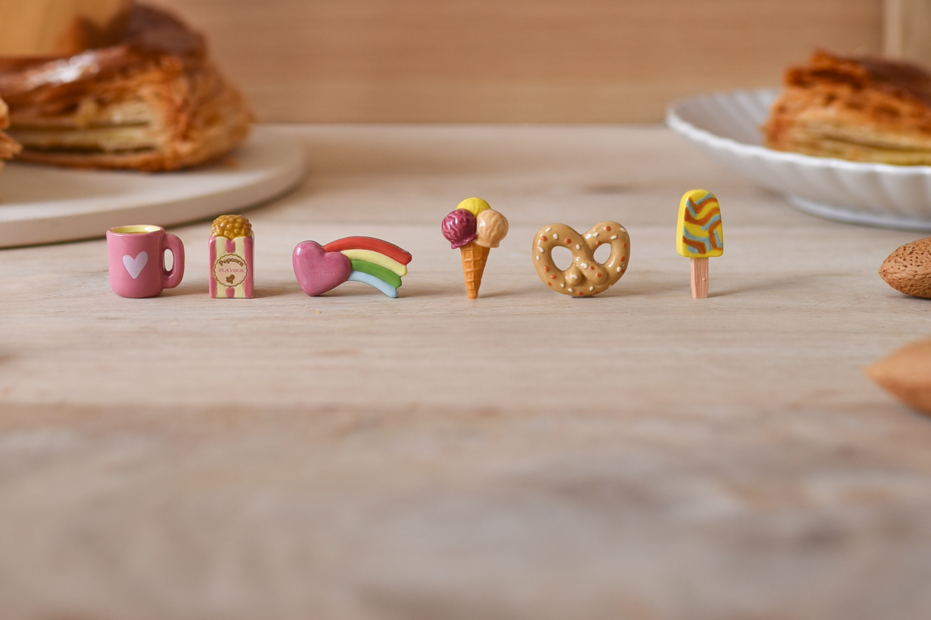 Six fèves en porcelaine alignées en fond une galette des rois. Les fèves dans l'ordre : un mug rose avec un coeur, un paquet de popcorn, un coeur rose avec un arc-en-ciel sur le côté, un cornet de glace 3 boules, un bretzel et un bâtonnet de glace.
